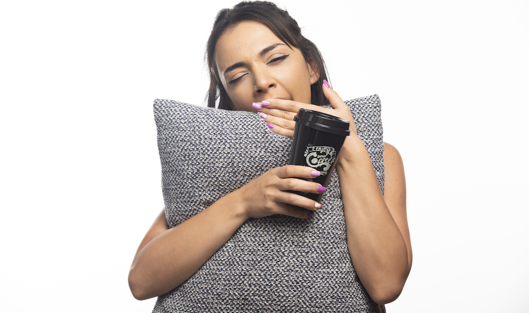 Woman yawning and holding a pillow with a cup on white background